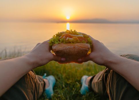 Unrecognizable young woman relaxing on coast by the sea with fast food fresh tasty burger and enjoying view of summer sunset, point of view.