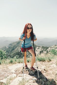 Sporty young woman in a t-shirt and shorts standing and resting with trekking poles in summer mountains outdoor.