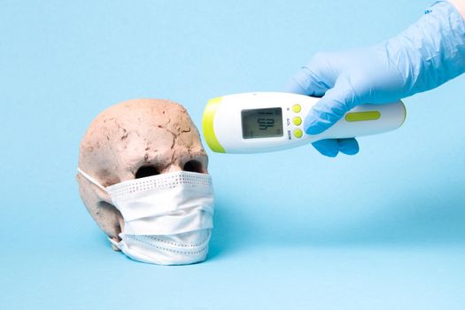 ceramic skull mask and a hand in a disposable medical rubber blue glove with infrared non-contact thermometer on a blue background, copy space, high temperature 40 degrees Celsius