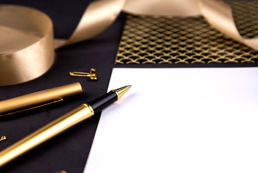 Gold pen, ribbon, paper clips and stationery on a black background with a white sheet of paper.