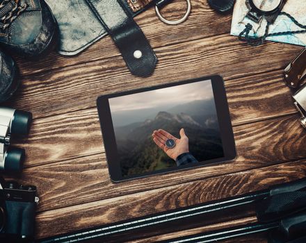 Equipment for travel and hiking on a wooden background, top view. Image with compass on a hand in mountains on screen of digital tablet.