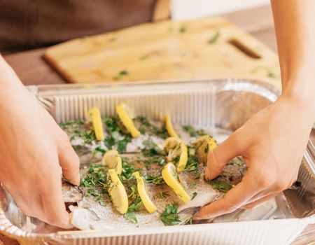 Unrecognizable young woman putting raw fish trout with lemons and greenery in baking foil dish in the kitchen.