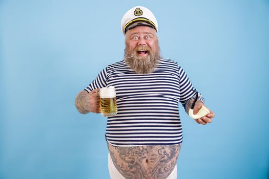 Joyful middle aged plus size person with beard in sailor suit holds smoking pipe and mug of fresh beer on light blue background in studio