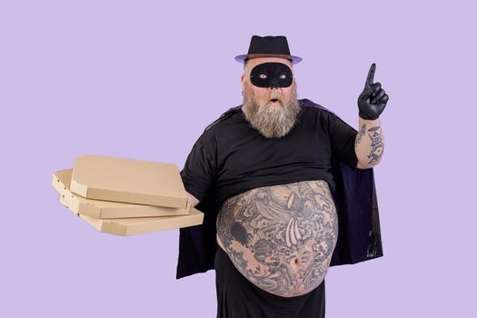 Surprised fat man in Zorro costume with large bare tummy holds cardboard boxes of pizza and points up standing on purple background in studio