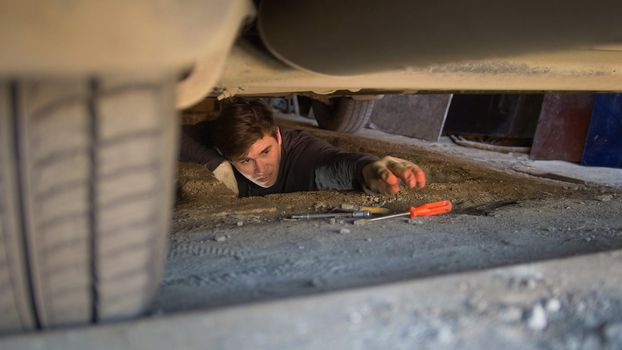 Man underneath a car reaching for a screwdriver for repairing car, close up