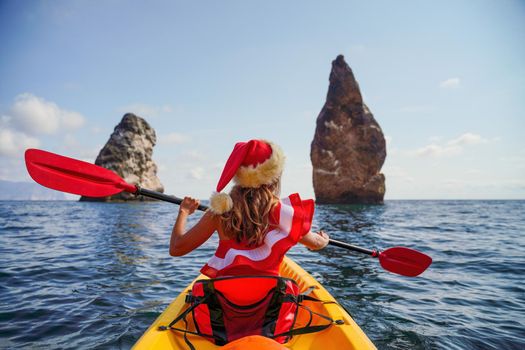 Young brunette woman in red swimsuit and Santa hat, swimming on kayak around basalt rocks like in Iceland. Back view. Christmas and travel concept