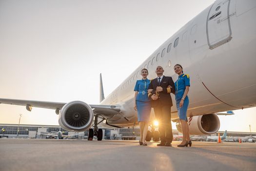 Full length shot of male pilot posing together with two female flight attendants in blue uniform in front of an airplane in terminal at sunset. Aircraft, aircrew concept