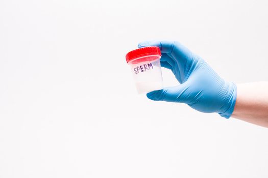 male hand in a blue disposable medical glove holds a jar for analysis with sperm, the inscription on the bank "sperm", white background, copy space, laboratory and analyzes concept