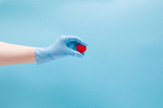 female hand in blue disposable medical glove holds a small red heart with a smile, smiling heart, health care concept, blue background, copy space, vari cordiologist treats