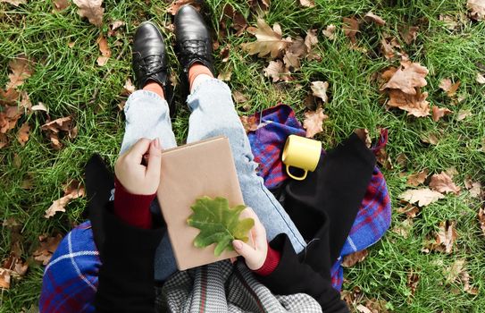 A woman holds a closed book lying on her lap with a fallen oak leaf in a park on a sunny warm autumn day on a green meadow. The concept of relaxation, reading and relaxation alone. Top view, flat lay