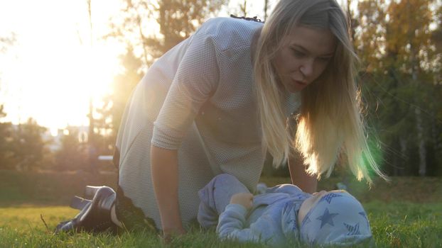 Young mother and her little baby playing in park. The baby lying on the grass. Close up. Wide shot