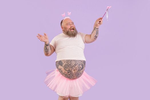 Expressive bearded mature man with overweight wearing fairy suit waves magic stick with butterfly posing on purple background in studio