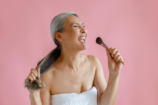 Jouful middle aged Asian woman sings song using cosmetic brush as microphone on pink background in studio. Mature beauty lifestyle