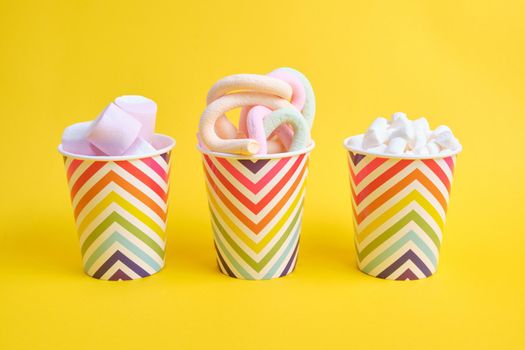 different types of marshmallows in festive paper cups with geometric pattern on yellow background, copy space