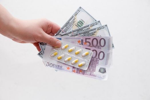 female hand holds a pack of yellow capsule pills and dollar bills euro and dollars, white background