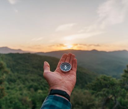 Magnetic compass on male palm hand in summer mountains at sunrise outdoor, point of view. Concept of travel and nature explore.