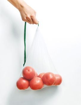 Female hand holding a reusable eco mesh bag with tomatoes on a white background. Zero waste concept.
