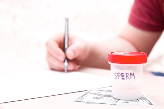 a test jar with a red cap and the inscription “Sperm” stands on dollar bills, against the background a man signs documents, a copy place, sperm in a jar, a sperm donor concept, sell sperm, close up