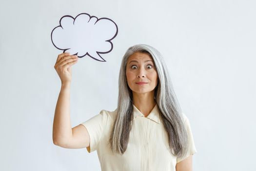 Surprised middle aged Asian woman with grey hair holds blank text bubble standing in light background in sudio. Mockup for design