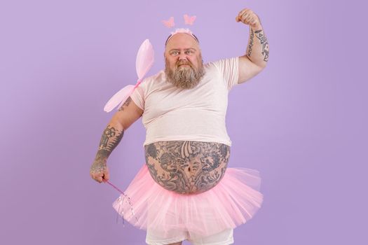 Funny brutal bearded obese man with tattoos wearing fairy costume with magic stick and wings shows muscles on purple background in studio