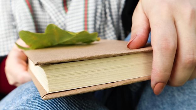 A woman holds a closed book lying in her lap with a fallen oak leaf close-up in a park on a sunny warm autumn day. The concept of relaxation, reading and relaxation alone