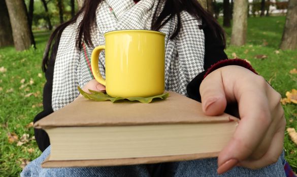 A girl dressed in a coat and a scarf in the autumn forest holds a book and a cup with a hot drink in her hands close-up in a city park on a warm day. The concept of reading, relaxation and comfort