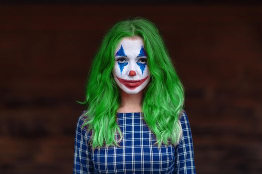 Close-up portrait of a greenhaired woman in chekered dress with joker makeup on a blurry brown background.