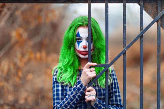 Close-up portrait of a greenhaired woman in chekered dress with joker makeup on a blurry brown background.