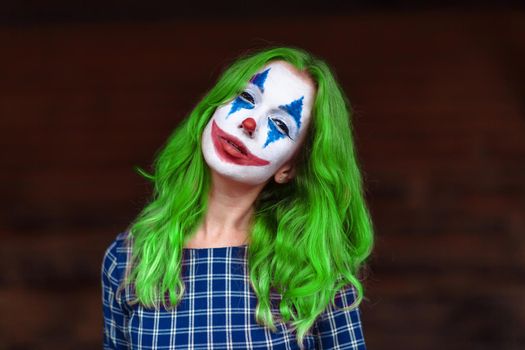 Close-up portrait of a greenhaired woman in chekered dress with joker makeup on a blurry brown background.