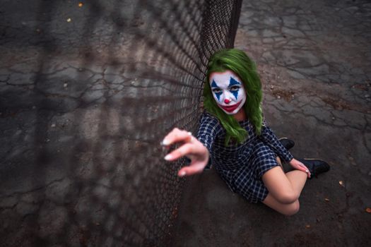 Greenhaired girl in chekered dress with joker makeup sitting near wire mesh fence on the ground. Wide shot.