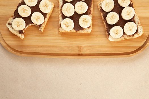 Three banana white bread toasts smeared with chocolate butter that lie on a cutting board on craft paper background. top view with area for text