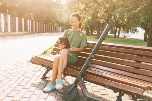 Beautiful young woman in casual green dress resting on wooden bench with digital tablet and an electric scooter in summer park.