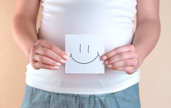A close-up view of the belly of a pregnant woman in a white T-shirt that is holding a piece of paper with a smiley face
