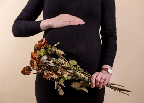 Side view of an attractive pregnant woman caressing her belly and holding a bouquet of dry roses in black dress while standing against yellow background.