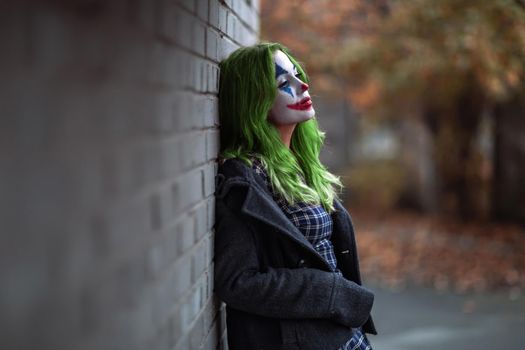 Portrait of a greenhaired girl in chekered dress with joker makeup on a brick wall blurred background.