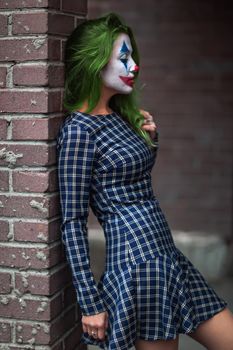 Portrait of a greenhaired girl in chekered dress with joker makeup on a brick wall blurred background.
