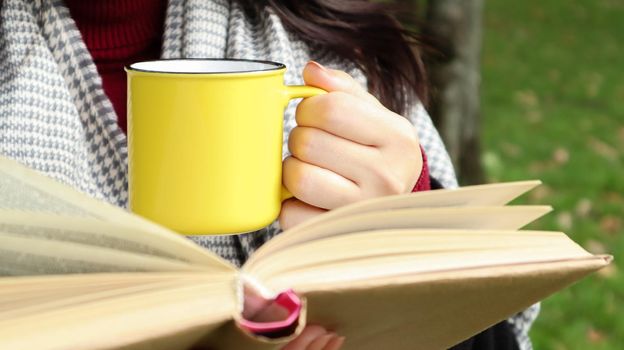 A girl dressed in a coat and a scarf in the autumn forest holds a book and a cup with a hot drink in her hands close-up in a city park on a warm day. The concept of reading, relaxation and comfort