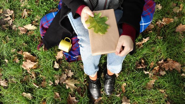 A woman holds a closed book lying on her lap with a fallen oak leaf in a park on a sunny warm autumn day on a green meadow. The concept of relaxation, reading and relaxation alone. Top view, flat lay