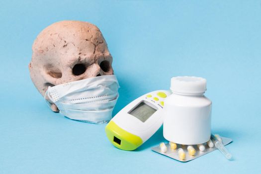 pills, drugs, syringe, infrared non-contact thermometer and ceramic skull in a protective medical face mask on a blue background, copy space, high temperature concept