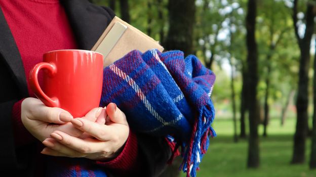 A red cup of coffee and a book with a blue checkered woolen blanket or plaid in the hands of a woman wearing a sweater and a black coat in the park. Warm and sunny weather. Soft cozy photography.