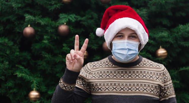 Christmas piece. Close up Portrait of man wearing a santa claus hat, xmas sweater and medical mask with emotion. Against the background of a Christmas tree. Coronavirus pandemic