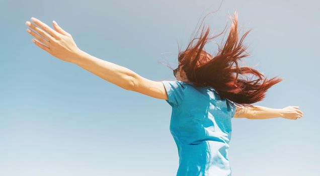 Happy active young woman standing with raised arms on background of blue sky in windy weather.