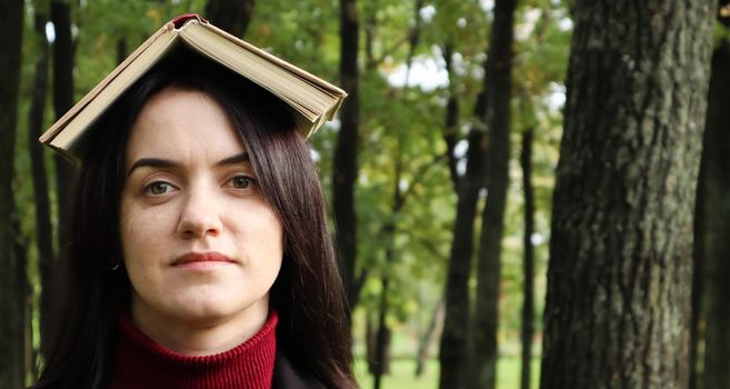 Portrait of a young and funny brunette in the park holding an open book on her head. Learning is fun. Woman balancing with books on her head. The student is tired of reading