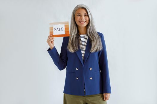 Pretty middle aged Asian lady with long grey hair wearing blue jacket holds card with word Sale posing on light background in studio