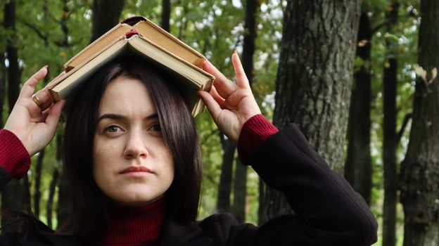 Portrait of a young and funny brunette in the park holding an open book on her head. Learning is fun. Woman balancing with books on her head. The student is tired of reading