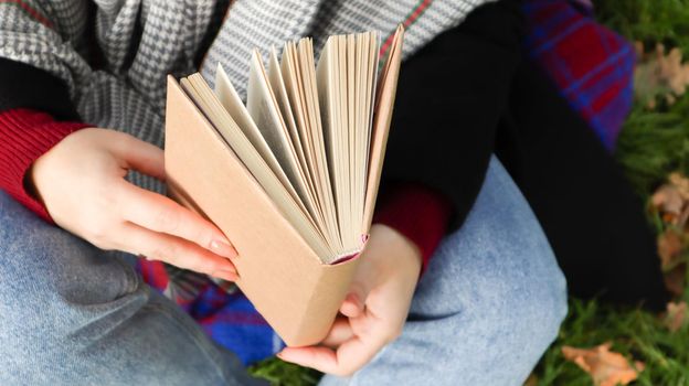 Girl reading a book in the autumn park. Female hands open the pages of a paper book outdoors on a warm sunny day. The student is preparing for the exam. Literary leisure in nature