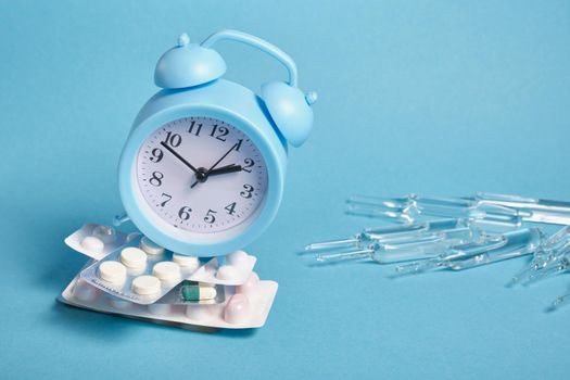 large ampoules, light blue alarm clock on a pile of packaging of different pills on a blue background, copy space, taking medications on time concept