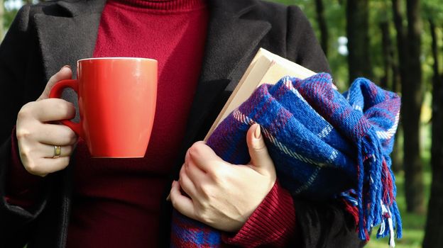 A red cup of coffee and a book with a blue checkered woolen blanket or plaid in the hands of a woman wearing a sweater and a black coat in the park. Warm and sunny weather. Soft cozy photography.