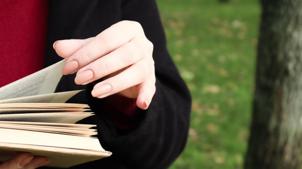 Girl reading a book in the park. Female hands flipping pages of paper book outdoors. The student is preparing for the exam. Literary leisure in nature. Close-up, copy space