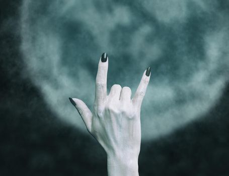 Hand of a dead zombie man with rock gesture in front of full moon. Halloween and horror concept.